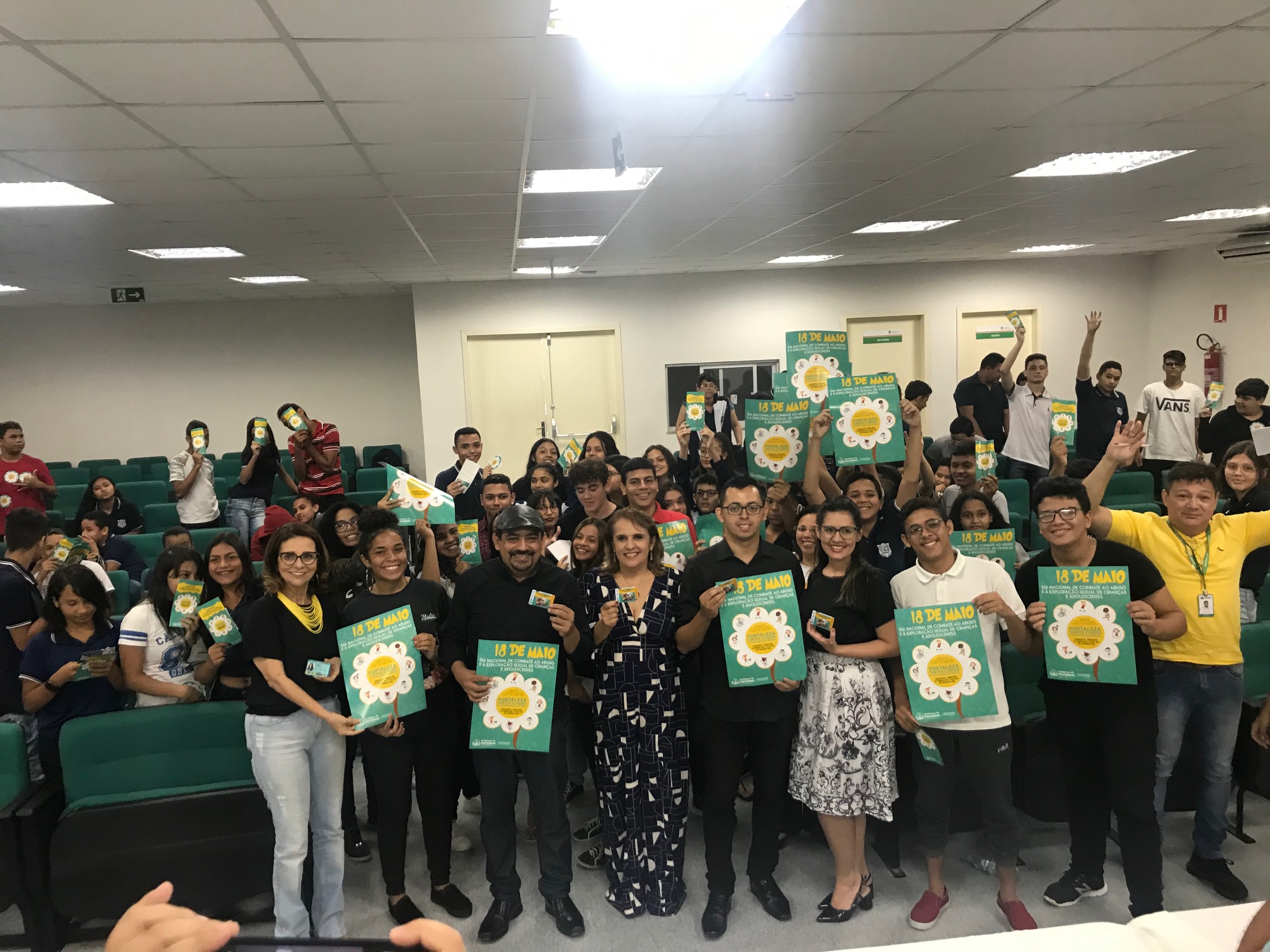 adultos e jovens em pé em auditório sorrindo e segurando panfletos e cartazes e posando para a foto